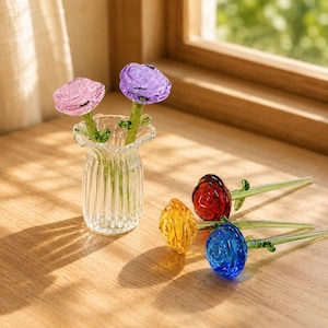 Può includere: Un vaso in vetro trasparente a coste contenente una rosa in vetro rosa e una viola, accompagnate da tre ulteriori rose in vetro rosso, giallo e blu, ciascuna dotata di uno stelo in vetro verde e un piccolo dettaglio a foglia su legno.