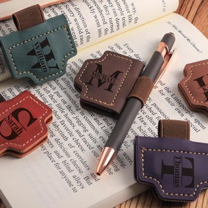 Puede incluir: Una colección de portalápices y marcadores de cuero personalizados en varios colores, incluyendo verde, marrón, rojo y morado. Cada uno presenta un nombre o inicial, y un bolígrafo está insertado en uno. Sobre un libro abierto.