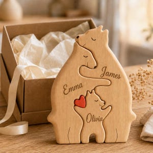 May include: A light-colored wooden puzzle featuring three interlocking bear shapes. The bears are engraved with the names Emma, James, and Olivia. A small, solid red heart is attached to the smallest bear figure. The puzzle stands upright.