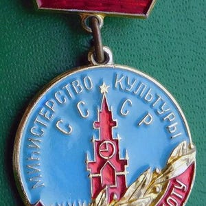 Könnte beinhalten: Vintage sowjetische Medaille mit rotem Emaille-Oberteil und rundem Anhänger. Der Anhänger hat einen blauen Hintergrund mit kyrillischem Text, ein rotes Gebäude und ein Banner mit Text. Wahrscheinlich eine Auszeichnung aus der UdSSR.
