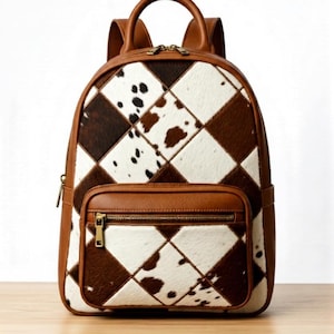 May include: A cowhide backpack featuring a diamond pattern in brown and white. It has brown leather detailing, a zipped front pocket, and a top handle. The brand name "SHAFHIDES" is displayed at the top.