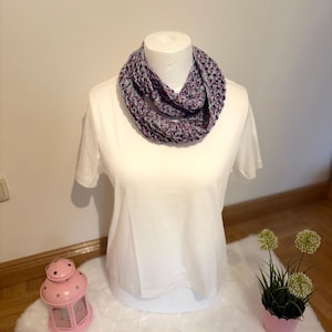 May include: A crocheted infinity scarf featuring a variegated yarn pattern in shades of purple, blue, and white. The scarf is displayed on a white mannequin torso wearing a plain white short-sleeved t-shirt, showcasing the textured loop design.