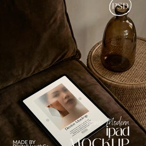 May include: An iPad mockup with a blurred image on the screen, placed on a brown velvet cushion. A brown glass bottle and a woven side table are in the background. The screen displays "Device Mockup" and "Organic Modern iPad Mockup".