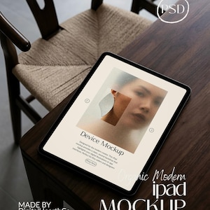 Pode incluir: Uma maquete de iPad preta com uma tela mostrando o rosto de uma mulher e formas abstratas. O iPad está em uma mesa de madeira escura ao lado de uma cadeira tecida. O texto na tela diz "Device Mockup" e "Organic Modern iPad Mockup".