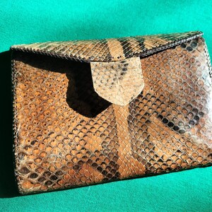 May include: A vintage snakeskin clutch bag with a flap closure. The bag displays a pattern of brown, tan, and black scales. The flap is secured with a small, light-coloured tab. The bag is rectangular and appears to be made of genuine snakeskin.