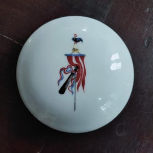 May include: A white, circular ceramic lid with a painted design. The design includes a sword, red and blue ribbons, and a rooster. The letters "RF" are visible. The lid is likely for a decorative container.