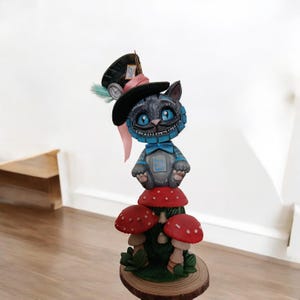 Op de afbeelding: Een grillige sculptuur van de Cheshire Cat uit Alice in Wonderland. De grijze kat met een brede grijns draagt een hoge hoed en een vlinderdas, zittend op rode paddenstoel figuren. De basis is een houten schijf.