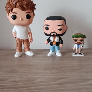 Può includere: Tre figure di Funko Pop sono esposte su una superficie di legno. Le figure includono un uomo in maglietta bianca e pantaloni marroni, un uomo in giacca nera e jeans blu e una piccola figura con una corona di foglie e occhiali.
