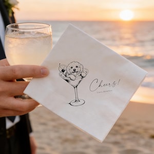 Puede incluir: Servilleta de cóctel blanca con una ilustración negra de un perro en una copa de martini, adornada con una cereza. El texto "Cheers!" y "love, Bentley" están escritos en la servilleta. Una persona sostiene la servilleta y un vaso de líquido.