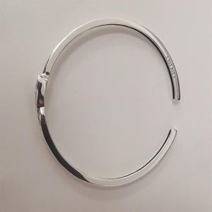 May include: A silver-colored hinged bangle bracelet. The bracelet is open, with the ends slightly separated. The inside of the bracelet is stamped with "S925 ALE 2". The bracelet has a smooth, polished finish and a simple, elegant design.
