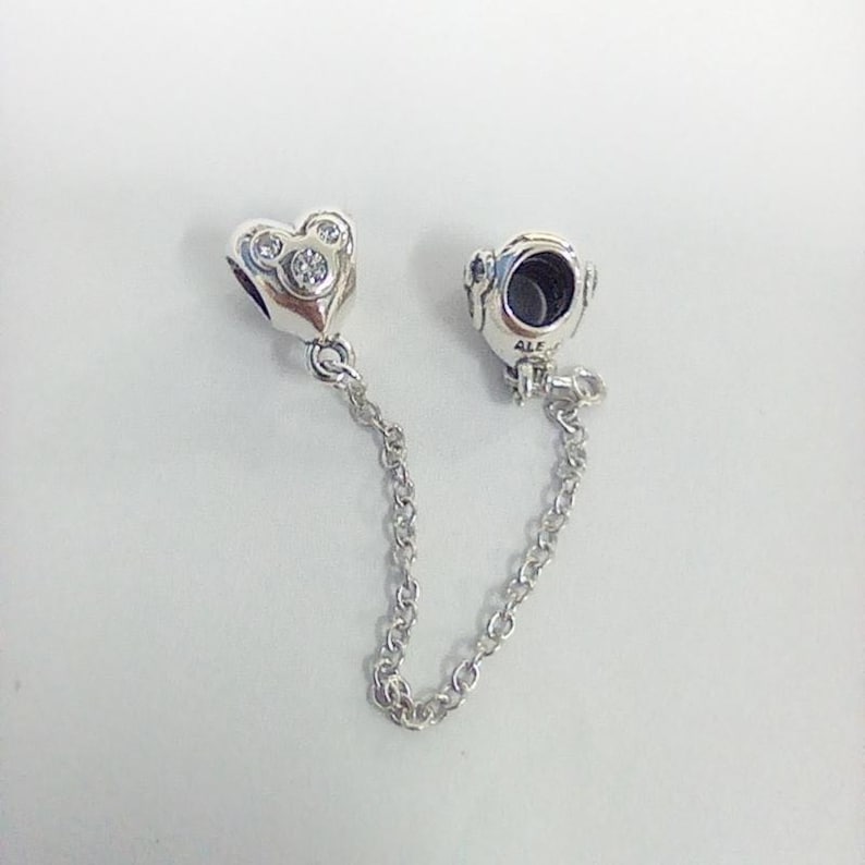 Op de afbeelding: Zilveren bedelarmband veiligheidsketting met een hartvormige bedel en een ronde bedel. De hartbedel heeft drie heldere stenen. De ketting is gemaakt van kleine, verbonden ringen.