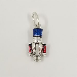 May include: A silver-colored charm in the shape of a toy soldier nutcracker. The charm has a blue hat, red accents, and the word "PANDORA" printed on the front. The charm is designed to be attached to a bracelet or necklace.