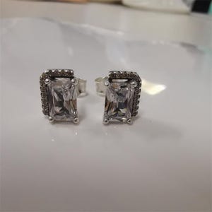 Könnte beinhalten: Ein Paar silberne Ohrstecker. Jeder Ohrring hat einen rechteckigen, klaren Edelstein, der von einem Kranz aus kleinen, dunklen Edelsteinen umgeben ist. Die Ohrringe liegen auf einer weißen Oberfläche.