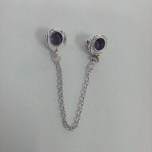 Op de afbeelding: Een zilveren veiligheidsketting voor een bedelarmband met twee ronde bedels. Elke bedel heeft een donker centrum en is verbonden door een fijne ketting. De bedels hebben een gedetailleerd ontwerp.