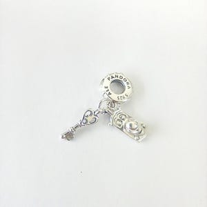 May include: A silver Pandora charm featuring a circular bail stamped with PANDORA, ALE, and S925. Two dangling pendants are attached to the bail: a small ornate key and a rectangular clock-shaped charm with a circular face and detailed markings.