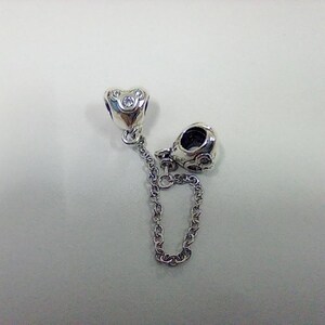 Op de afbeelding: Een zilveren bedel met een hartvormige kraal en een ronde kraal, verbonden door een ketting. De hartkraal heeft een kleine heldere steen in het midden. De bedel is voor een armband of ketting.