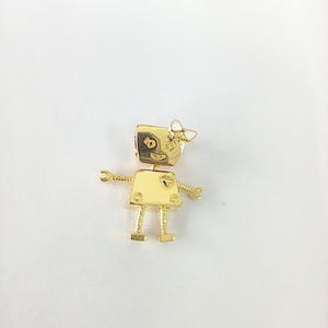 May include: A gold-colored robot-shaped brooch with a bow on its head. The robot has a square head with a face and a rectangular body with arms and legs. The brooch is made of a shiny metal and has a detailed design.