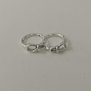 May include: Two silver-colored hoop earrings. Each earring features a bow-shaped design at the bottom. The earrings are displayed on a light gray surface.