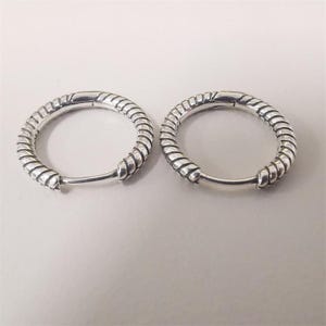 May include: A pair of silver hoop earrings. Each earring features a textured, twisted rope design around the hoop. The earrings are displayed against a white backdrop.