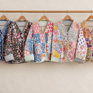 May include: Five quilted jackets with V-necklines hanging on a wooden rod. Each jacket features a unique patchwork design with floral patterns in various colors, including blue, black, pink, and yellow, with striped inner linings and cuffs.
