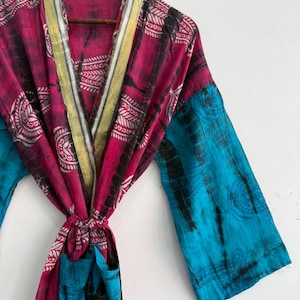 May include: A long-sleeved robe featuring a vibrant magenta and bright blue tie-dye pattern with white leaf-like motifs. The garment includes a wide, metallic gold-toned trim along the front opening and a matching fabric tie belt at the waist.