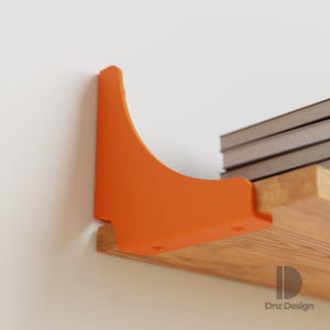 Op de afbeelding: Een oranje metalen plankbeugel is gemonteerd op een houten plank en ondersteunt een stapel boeken. De beugel heeft een gebogen ontwerp en is aan de muur bevestigd. De plank is van hout. Het ontwerp is van Dnz Design.