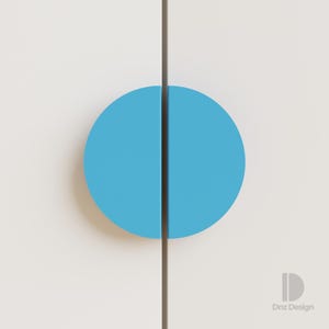 以下が含まれることがあります： 明るい青色の半円形の取っ手が付いた、白いキャビネットドアのクローズアップ。取っ手は、垂直の濃い茶色の線で半分に分割されています。キャビネットドアは白です。デザインはミニマリストでモダンです。