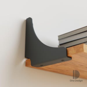 Puede incluir: Un soporte de estante de metal negro está montado en una pared blanca, soportando un estante de madera. El estante contiene una pila de libros. El soporte tiene un diseño curvo y está fijado al estante con tornillos.