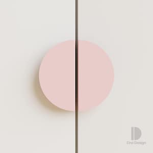 May include: A close-up of a cabinet door with a pale pink, semi-circular handle. The handle is bisected by a dark brown vertical line. The cabinet door is white. The design is minimalist and modern.