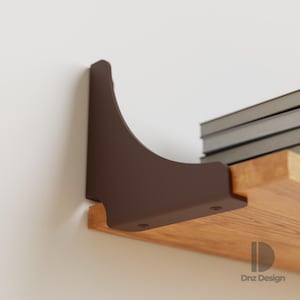 Puede incluir: Un soporte de estante de metal marrón fijado a una pared blanca, que sostiene un estante de madera con libros. El soporte tiene un diseño curvo y está asegurado con tornillos. El estante es de madera clara.