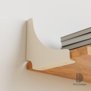 May include: A cream-coloured shelf bracket is mounted on a white wall, supporting a wooden shelf. The shelf holds a stack of books. The bracket has a curved design. The logo "Dnz Design" is visible.