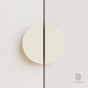 May include: A close-up of a cabinet door featuring a cream-coloured, semi-circular handle. The handle is divided by a dark vertical line, and the door is a light cream colour. The design is minimalist and modern.