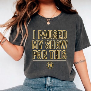 Puede incluir: Camiseta gris oscuro con el texto amarillo "I PAUSED MY SHOW FOR THIS" y un símbolo de reproducción. Esta camiseta informal es adecuada para el uso diario.
