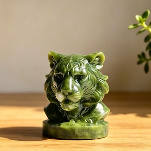 Könnte beinhalten: Eine geschnitzte Tigerkopf-Figur aus grünem Stein mit detaillierten Gesichtszügen, spitzen Ohren und einer glatten, polierten Oberfläche. Die Skulptur ist auf einem runden Sockel montiert und zeigt tiefe olivgrüne Töne sowie natürliche Texturen.