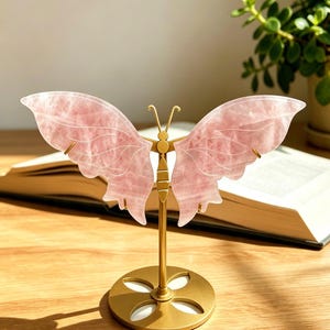May include: A decorative butterfly sculpture featuring two translucent, pale pink rose quartz wings attached to a central gold-toned metal body. The sculpture is supported by a thin vertical gold-toned metal rod and a circular gold-toned base.