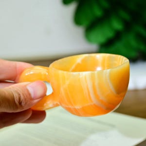 Könnte beinhalten: Eine kleine, handgeschnitzte Tasse mit Henkel aus orangefarbenem und weißem Wirbelstein. Die Tasse wird von einer Person in der Hand gehalten, im Hintergrund ein Buch. Die Tasse ist ein einzigartiges Trinkgefäß.