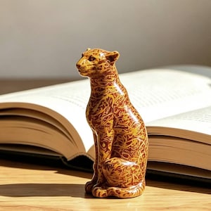 以下が含まれることがあります： ゴールデンブラウンとダークアンバーの色合いで、光沢のあるマーブル仕上げが施された小さなヒョウの置物。滑らかで反射する表面と詳細な顔の造形が特徴で、動物が直立した姿勢で座っている様子を捉えています。