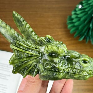 May include: A green dragon head sculpture, carved from a jade-like stone. The dragon features detailed scales, horns, and a defined snout. The sculpture is held in a hand, highlighting the craftsmanship and the dragon's features.