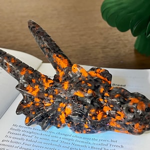 May include: A dragon head sculpture in a speckled pattern of black, grey, and orange. The sculpture rests on a book with visible text. The dragon has two horns and a detailed, textured surface. A decorative item.