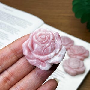 May include: A close-up of a pink rose-shaped stone carving. The stone has a light pink hue with white accents, and the rose petals are intricately detailed. Several other rose carvings are visible on a white surface.