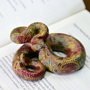 Puede incluir: Una detallada y colorida figura de serpiente con una superficie texturizada. La serpiente está enrollada y presenta tonos de rojo, amarillo, verde y marrón. Descansa sobre un libro abierto con texto visible.