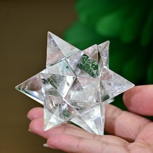 Puede incluir: Una estrella de cristal clara y multifacética, sostenida en una mano. La estrella tiene puntas triangulares afiladas y un diseño geométrico complejo. El cristal es transparente, permitiendo el paso de la luz, y parece ser un objeto decorativo o una herramienta espiritual.