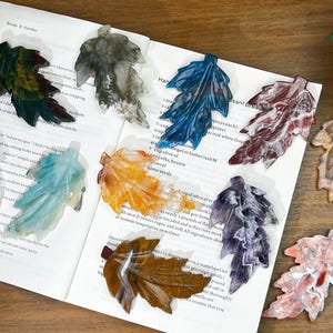 Puede incluir: Conjunto de adornos decorativos con forma de hoja en varios colores, incluyendo verde, azul y naranja. Estos objetos están colocados sobre un libro abierto con texto. Las hojas parecen ser de piedra.
