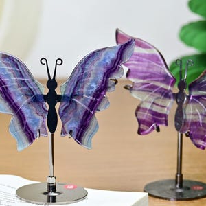 Puede incluir: Dos esculturas decorativas de mariposas. Las alas son de piedra translúcida morada y azul, con un patrón de rayas. Los cuerpos de las mariposas son negros y están montados sobre soportes de metal plateado. Las mariposas se exhiben sobre una superficie de madera.