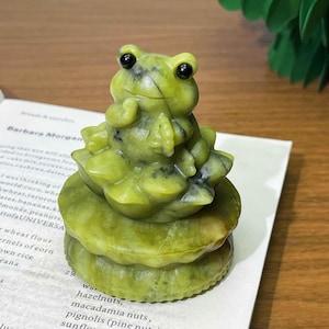 May include: A green frog figurine with black eyes, perched atop a layered base. The frog is carved from a light green stone, with darker green veining. The figurine is placed on an open book with text.
