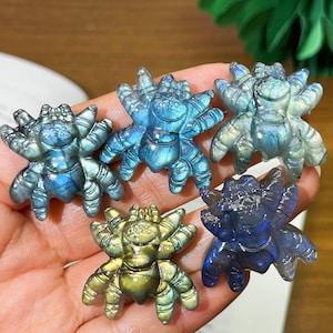May include: Five small spider figurines carved from labradorite stone. Each piece displays a unique iridescent sheen with flashes of blue, gold, and green light reflecting off the polished surfaces. The carvings feature detailed legs and bodies.