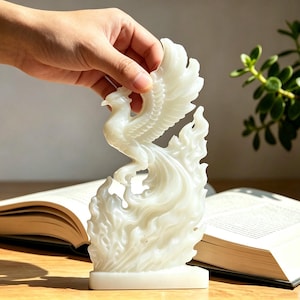 May include: A white, translucent phoenix figurine standing on a rectangular base. The sculpture depicts a bird with wings spread upwards, emerging from stylised, flame-like patterns that curve around the body and tail, creating a dynamic form.