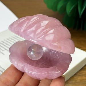 May include: A carved rose quartz clam shell with a pearl. The pink shell is open, revealing a sphere inside. The shell is resting on a book, with a green decorative object in the background.