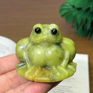 May include: A carved frog figurine in shades of green and yellow. The frog has large, black eyes and is sitting with its legs folded. The figurine is made of a smooth, polished stone and is a decorative item.