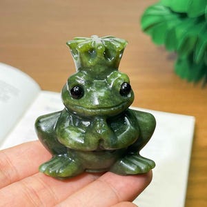 May include: A green jade frog figurine with a crown. The frog has black eyes and is posed with its hands together. The figurine is sitting on a book. The frog is a decorative item.
